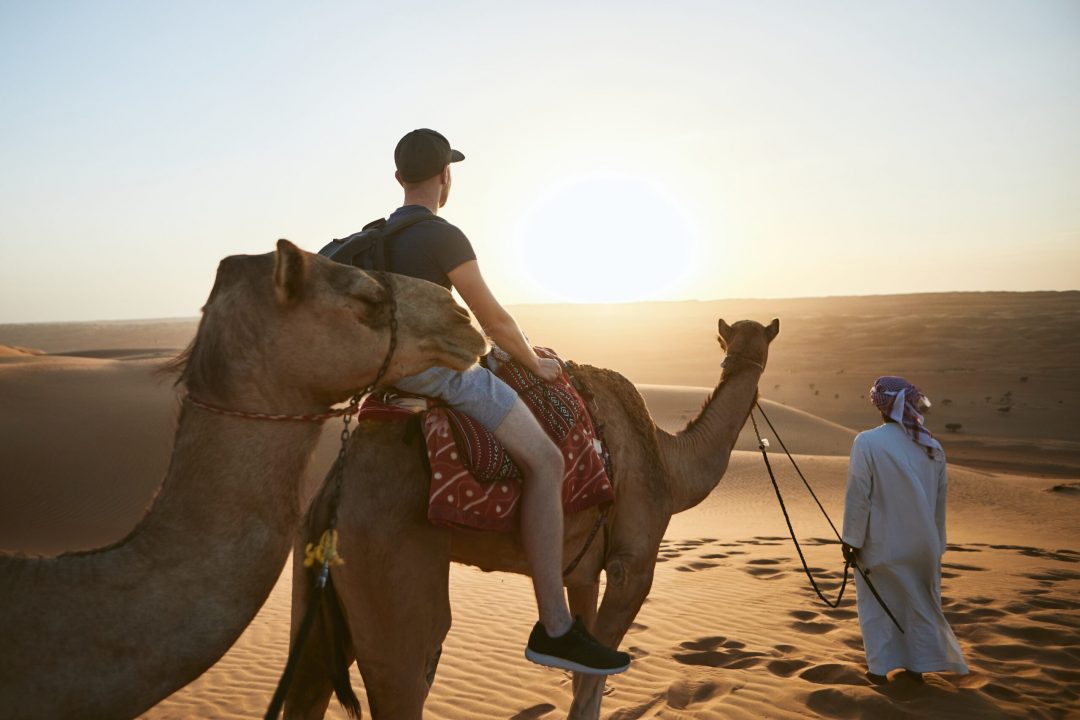 Camel riding in desert at amazing sunset. Young man enjoying journey on sand dunes. Wahiba Sands in Sultanate of Oman